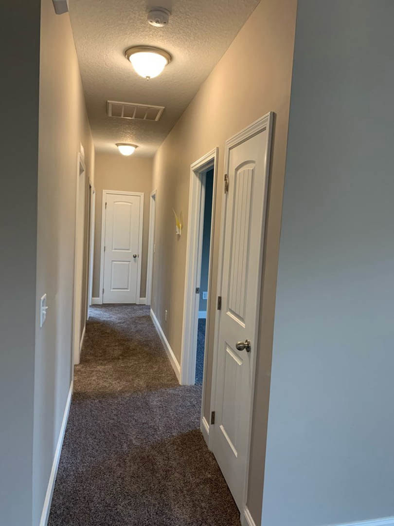Hallway with light beige carpet, multiple white paneled doors with silver knobs, white walls, and a ceiling-mounted light fixture