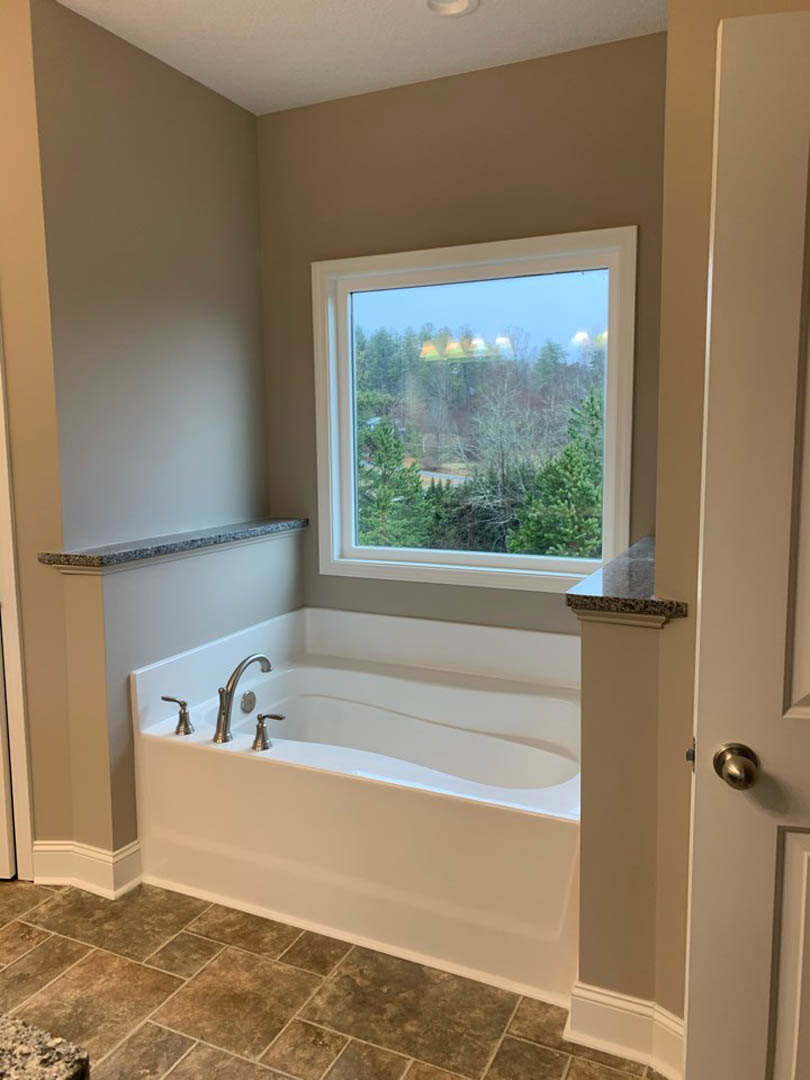 Freestanding bathtub with chrome faucet on light tile floor, large window overlooking trees, white walls, and modern door hardware