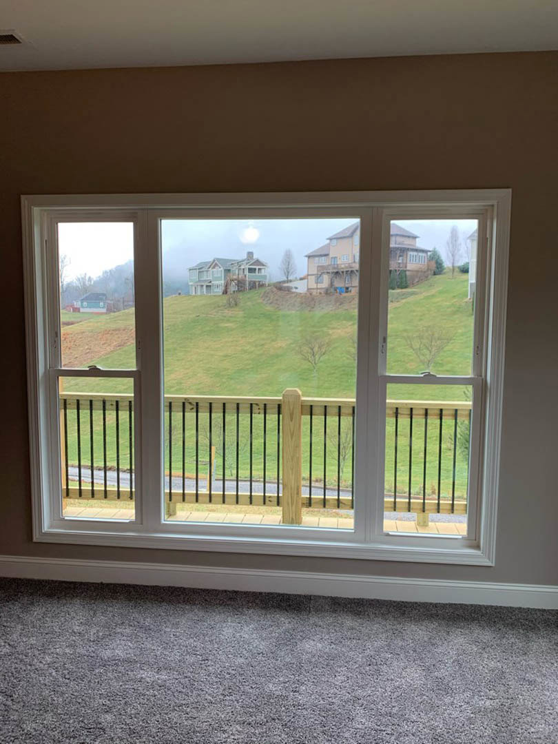 Large window framed by white trim, overlooking a grassy hill and wooden fence; beige carpet flooring and partial view of interior wall.