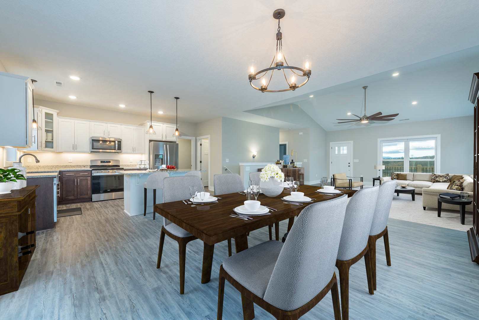 Wood dining table set with plates, glasses, and a white cup, surrounded by chairs in a modern dining room; overhead wire-wrapped light fixture, white vase with flowers, neutral
