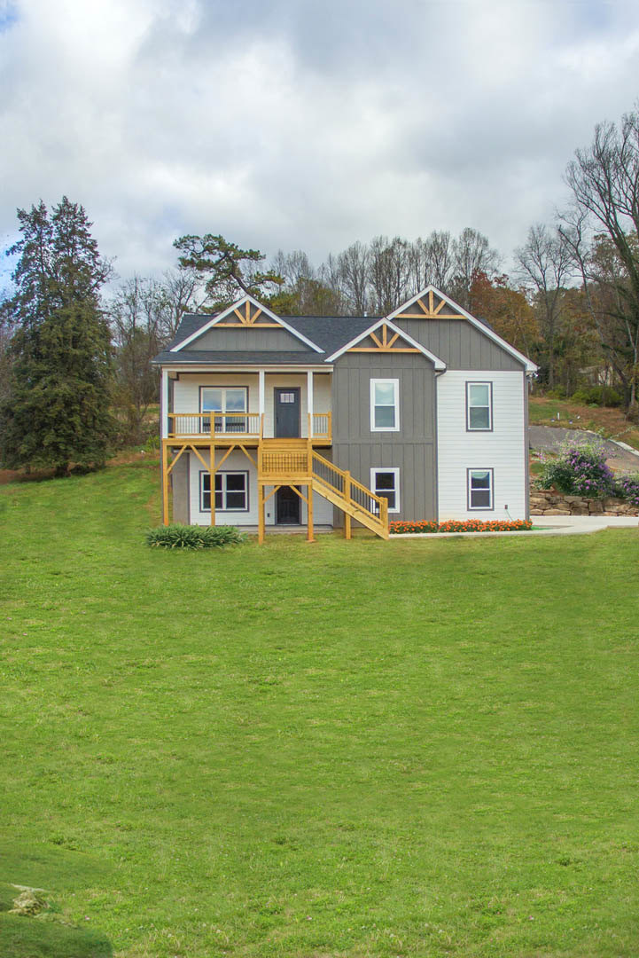 Two-story house with white siding, covered porch, wooden stairs, and spacious front lawn bordered by mature leafy trees.