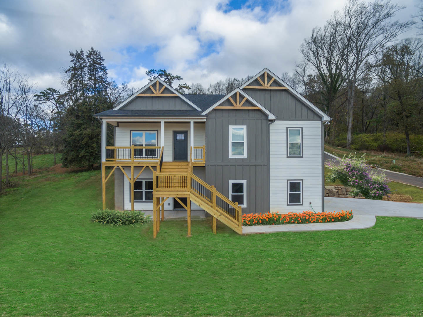 Modern cottage with wooden deck, black door with glass window, white-framed windows, wooden exterior stairs, lush green lawn, and cluster of orange flowers in front yard