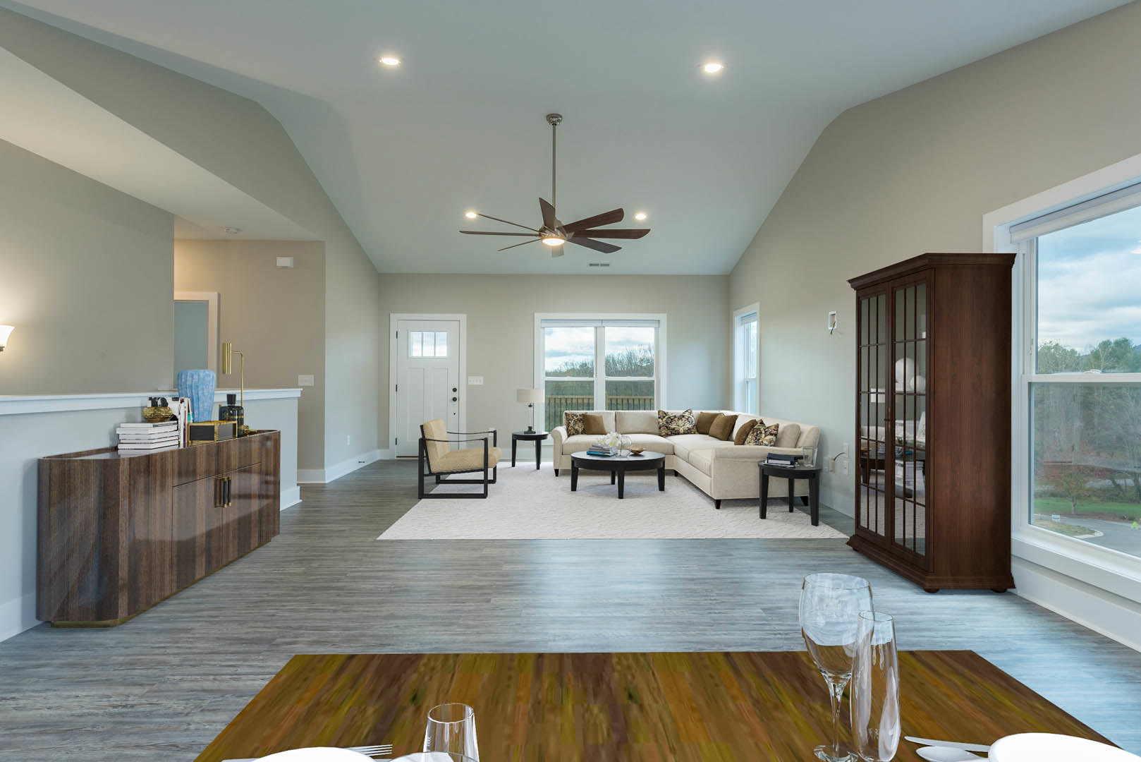 Living room with light-colored couch and wooden coffee table, ceiling fan with lights, black-framed chair, wooden cabinet with shelf, glass-front cabinet, laminate flooring