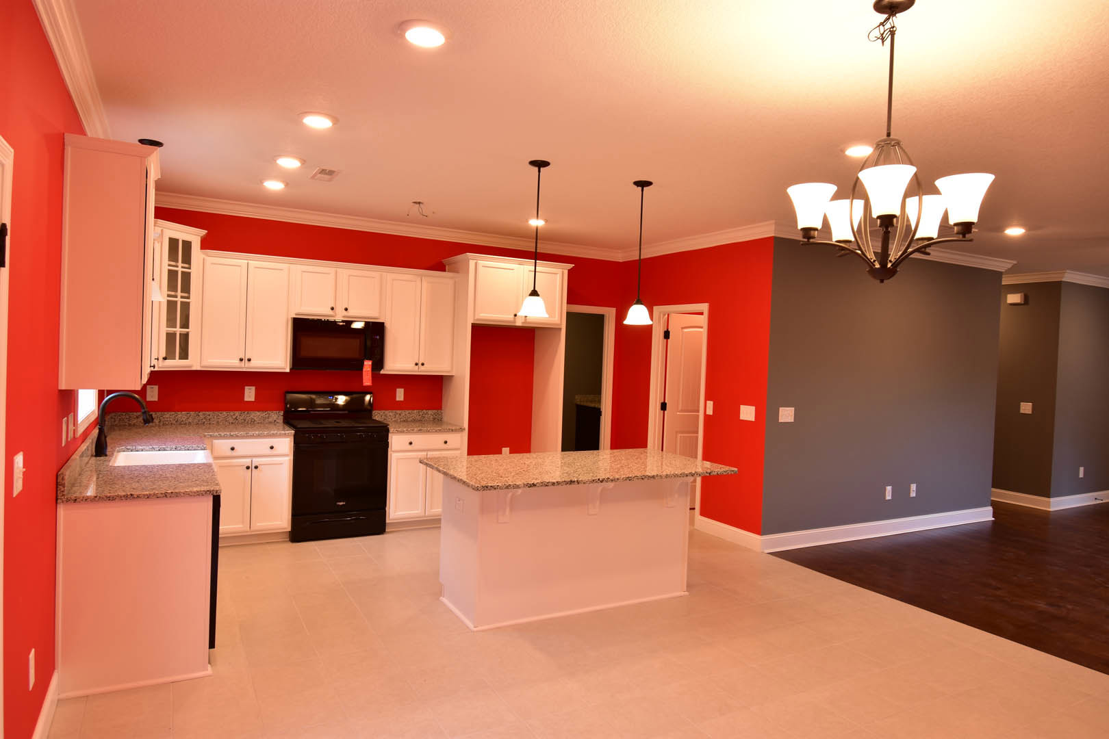 Modern kitchen featuring a central island with white countertop and base, black stove, built-in sink, cabinetry, chandelier light fixture, and TV screen mounted on the wall.