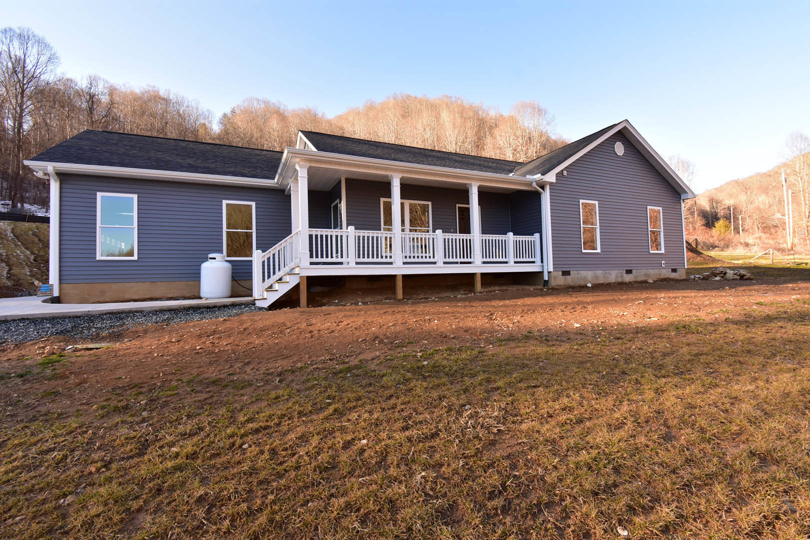 Two-story house with white siding, covered porch featuring white railings and stairs, large grassy yard, white-framed windows, and mature trees in the background