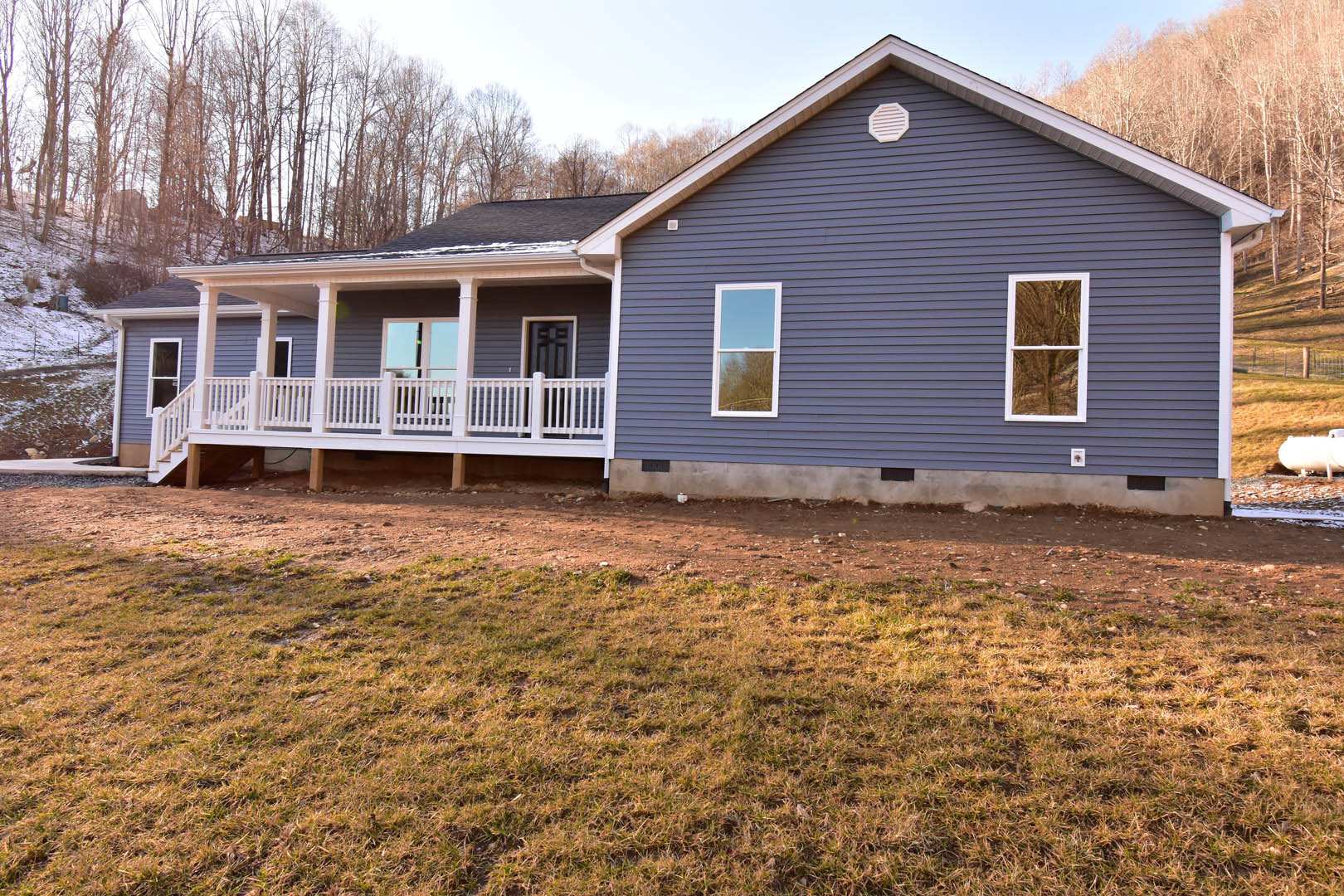 Two-story house with light siding, white-framed windows, covered porch with white railing, grassy front yard, concrete retaining wall, blue sky overhead