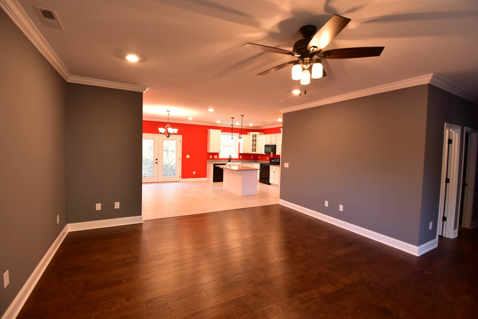 Open living room with hardwood floors, ceiling fan with lights, white kitchen island topped with marble, glass-paneled double doors, and plaster walls.