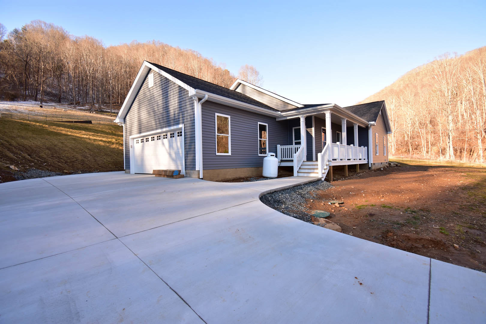 Two-story house with white siding, covered porch with white railing, concrete driveway, and grassy hill in the background.