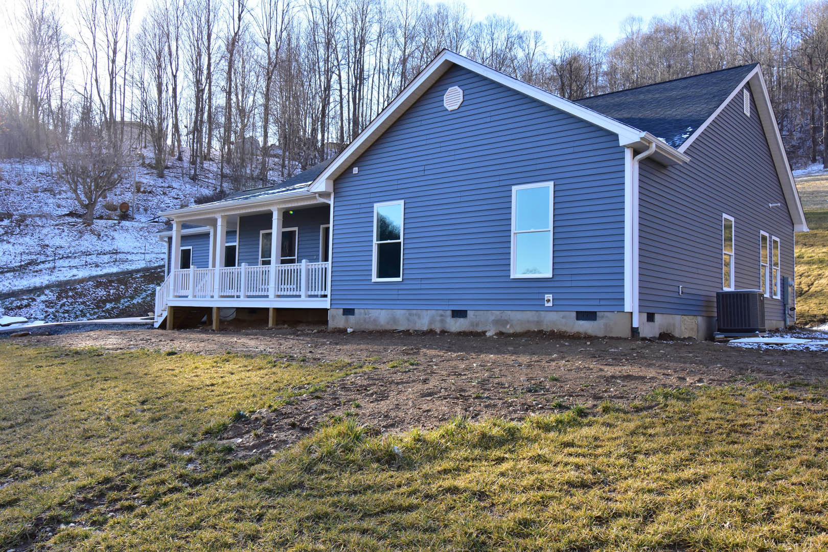 Blue siding house with white trim and porch, white railing, grassy hill in front, white-framed windows, tree branches visible against sky.