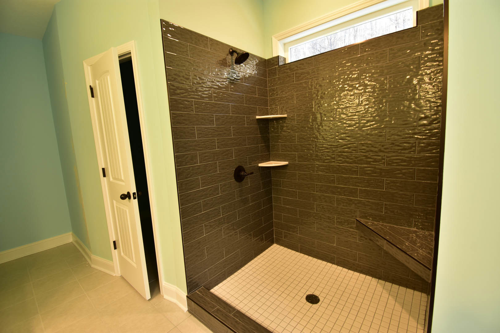 Walk-in shower with built-in shelf, wall-mounted shower head, and adjacent window; white tile walls, tiled floor with drain, modern sink nearby