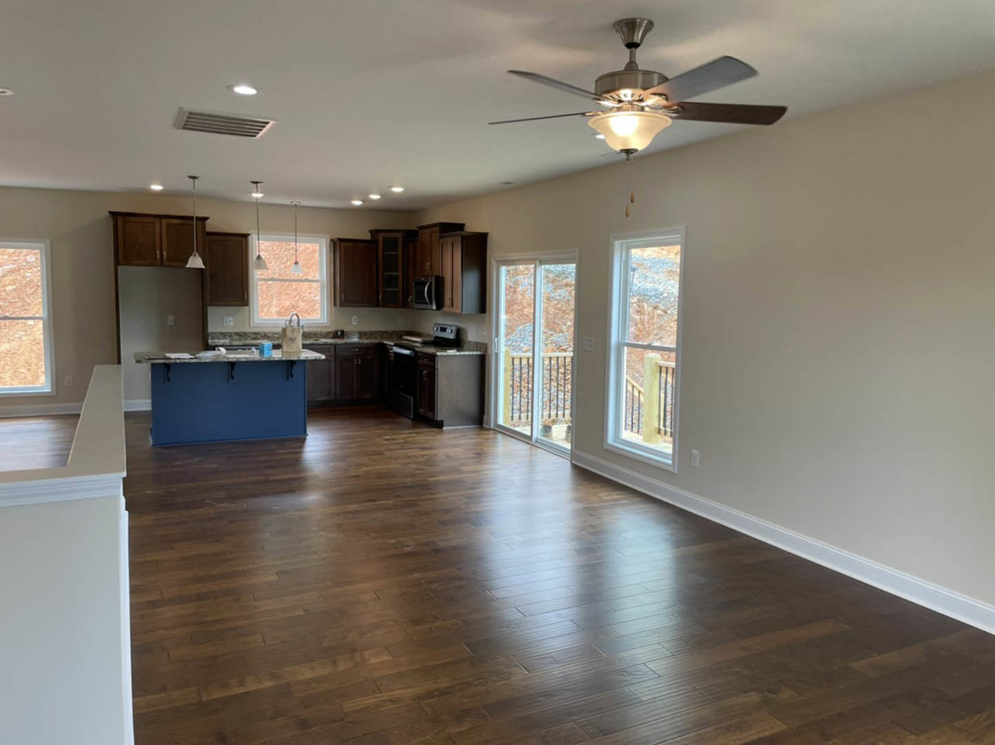 Open kitchen and dining area with wood cabinetry, stainless appliances, hardwood flooring, ceiling fan with light, and large window framed in white overlooking outdoor railing.