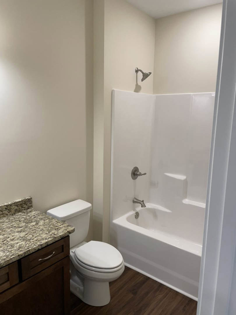 White toilet beside a marble countertop with black veining, white bathtub with silver faucets, light gray tiled walls, and shadow of exposed pipe on wall.