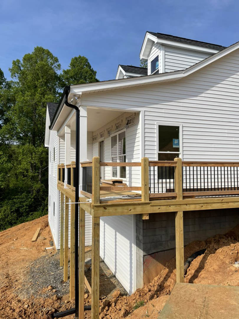 Partially built white house with black gutters, exposed wood deck and railing, window with construction sign, dirt and gravel yard.