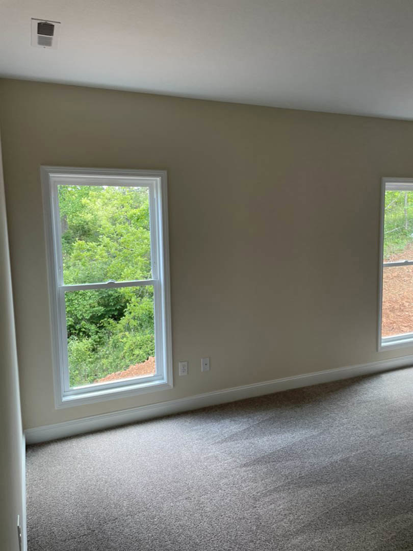 Carpeted bedroom with two large windows, white walls, and views of trees and grass outside