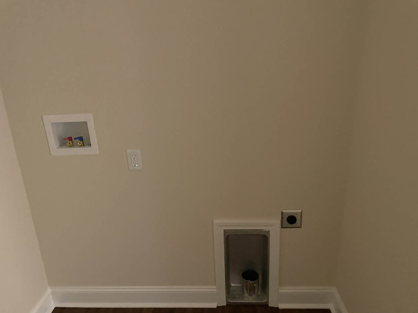 White plaster bathroom wall with a recessed metal trash can, white framed access panel featuring red and blue valve, white electrical outlet, and black circular fixture above