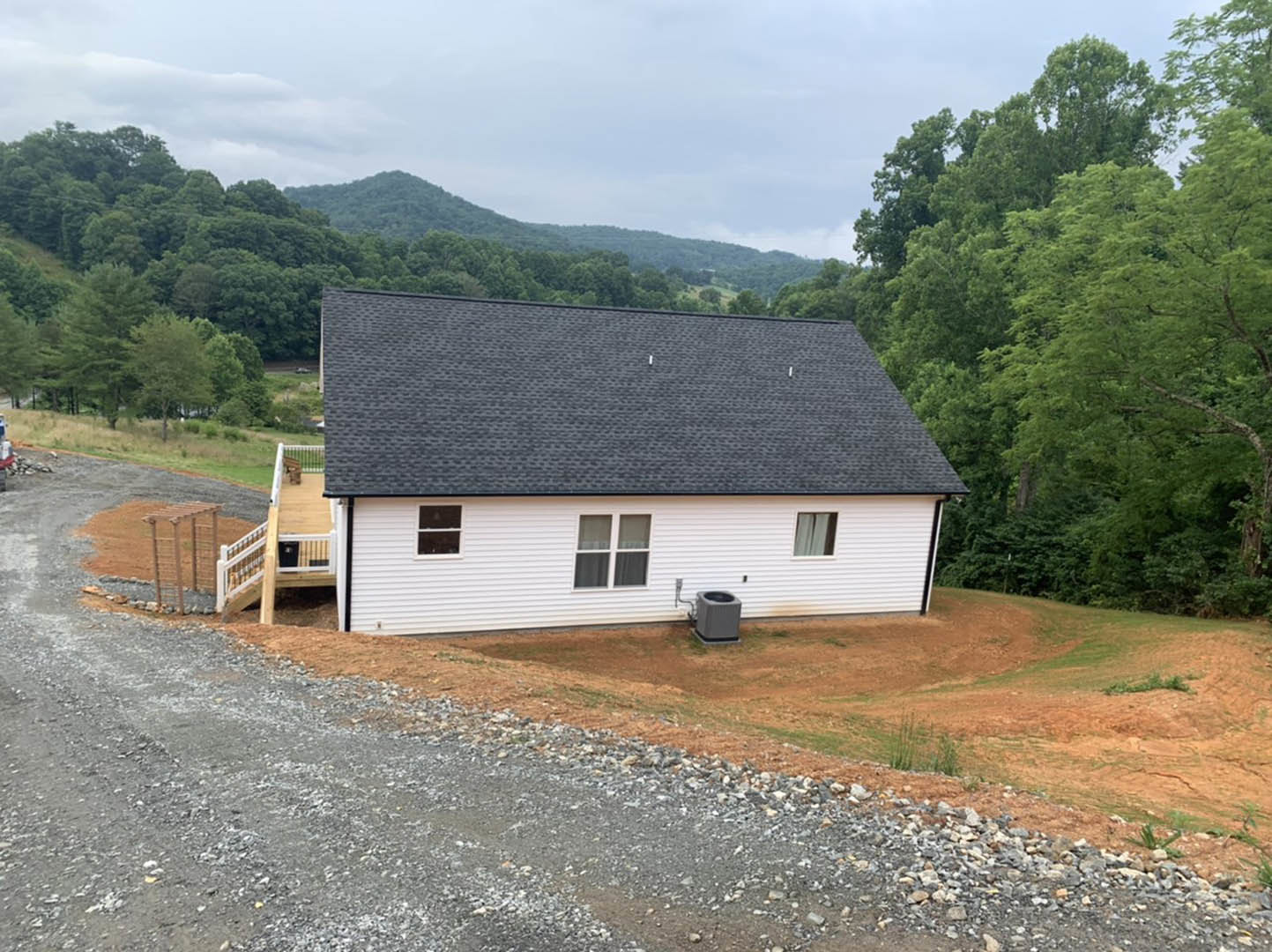 White house with black roof, front staircase leading to entry, gravel driveway, grassy yard, trees in background, white-framed windows with curtains visible.