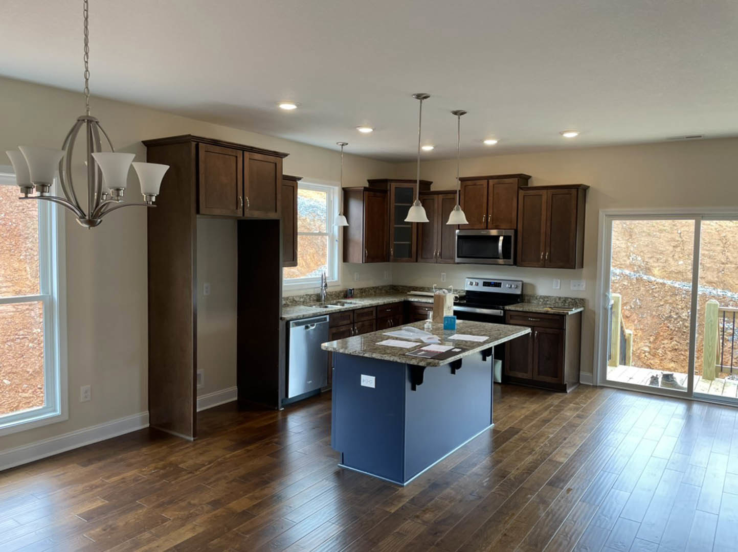 Open kitchen with wood flooring, central island topped with papers, white cabinetry, stainless steel microwave, sink, bell-shaped pendant light, and sliding glass door revealing