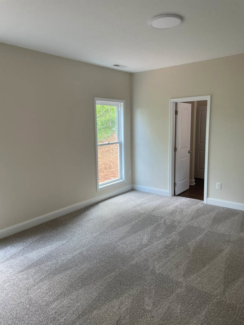 Bedroom with beige carpet, white paneled door with silver handle, white framed window, and white walls; partial view of bathroom with white toilet visible through open doorway.