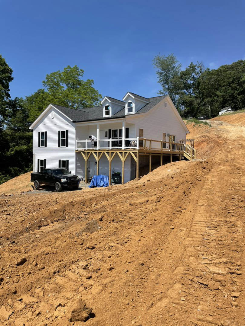 White house under construction on a hill with exposed framing, large front porch, white railing, blue tarp covering materials, black truck parked on dirt driveway, surrounded by