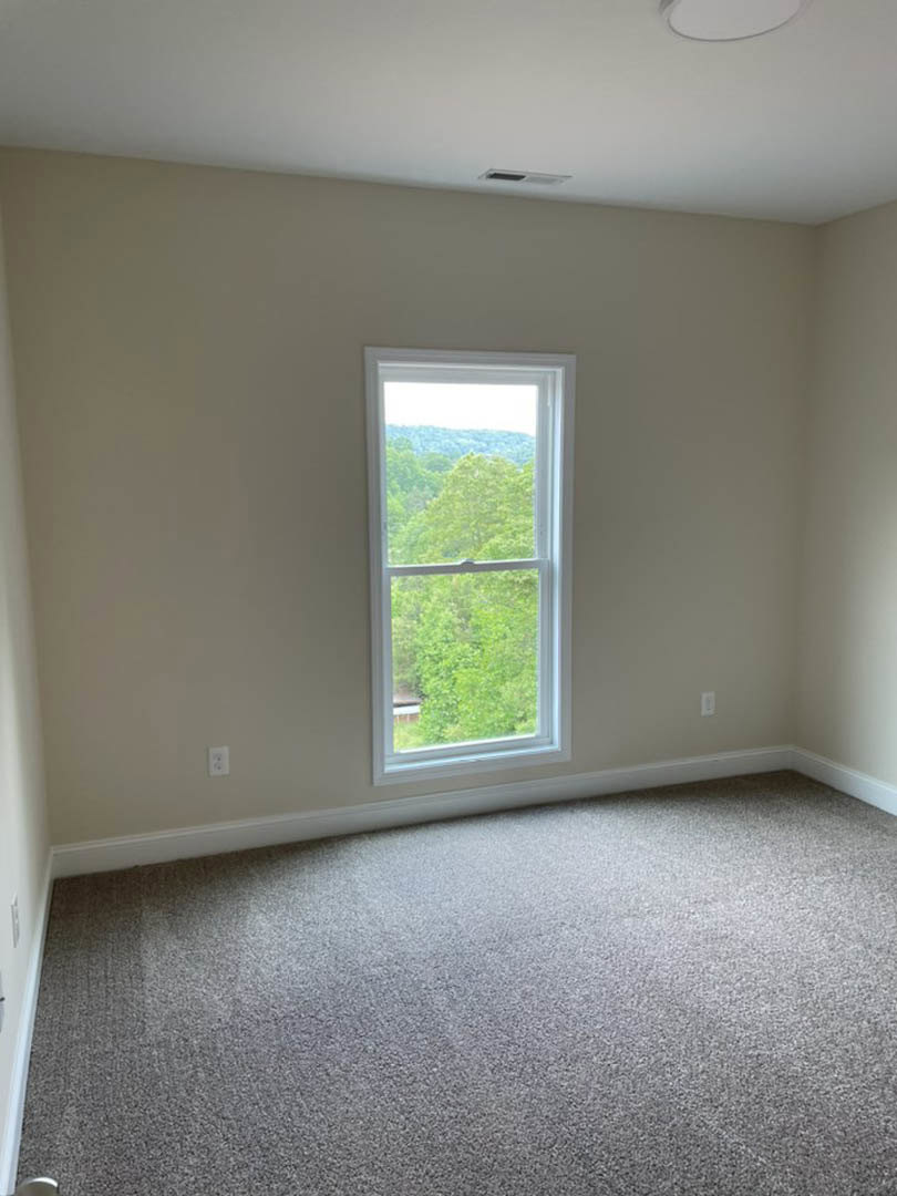Carpeted bedroom with large window overlooking leafy trees, white walls, and ceiling vent