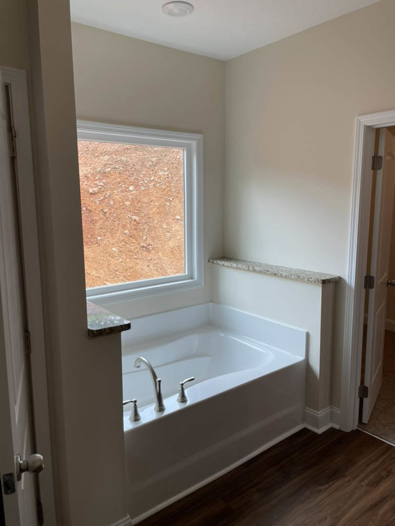 Freestanding white bathtub with chrome faucet beside a large window, surrounded by light gray tile walls and a stone countertop