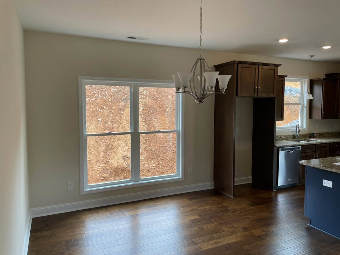 Kitchen with wooden flooring, white cabinetry, stainless steel sink beneath a window overlooking a dirt field, shadow cast on wall, modern chandelier overhead