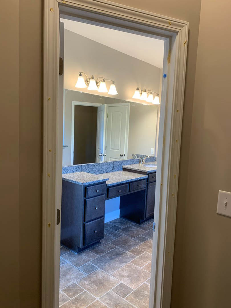 Bathroom with rectangular mirror above white vanity, three-light fixture, tile flooring, cabinetry with drawers, chrome faucet, and wall-mounted light switch.