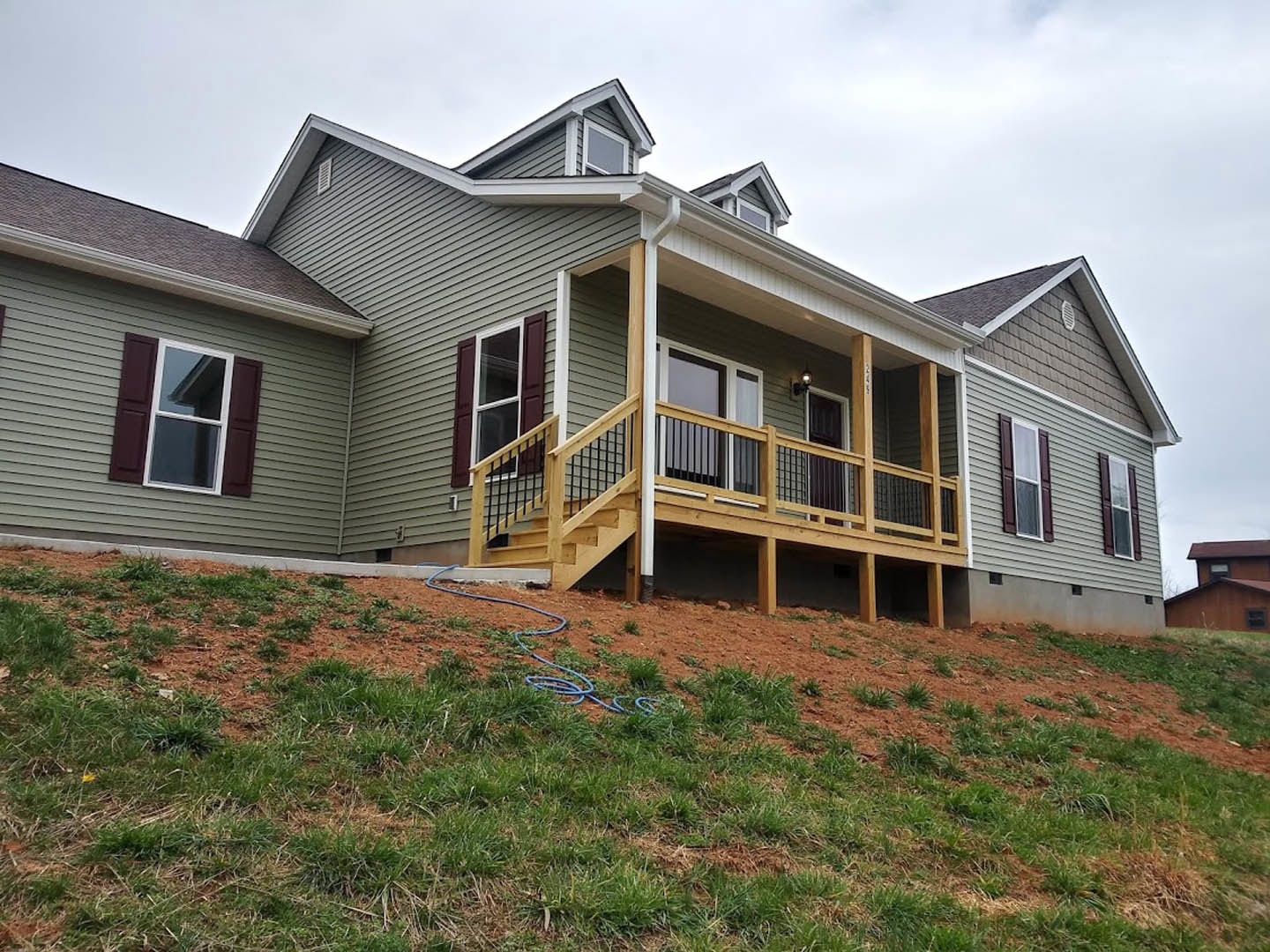 Two-story house with white siding, covered porch with brown railings, stairs leading to lawn, blue garden hose on grass, white-framed windows, sloped yard, and clear sky.