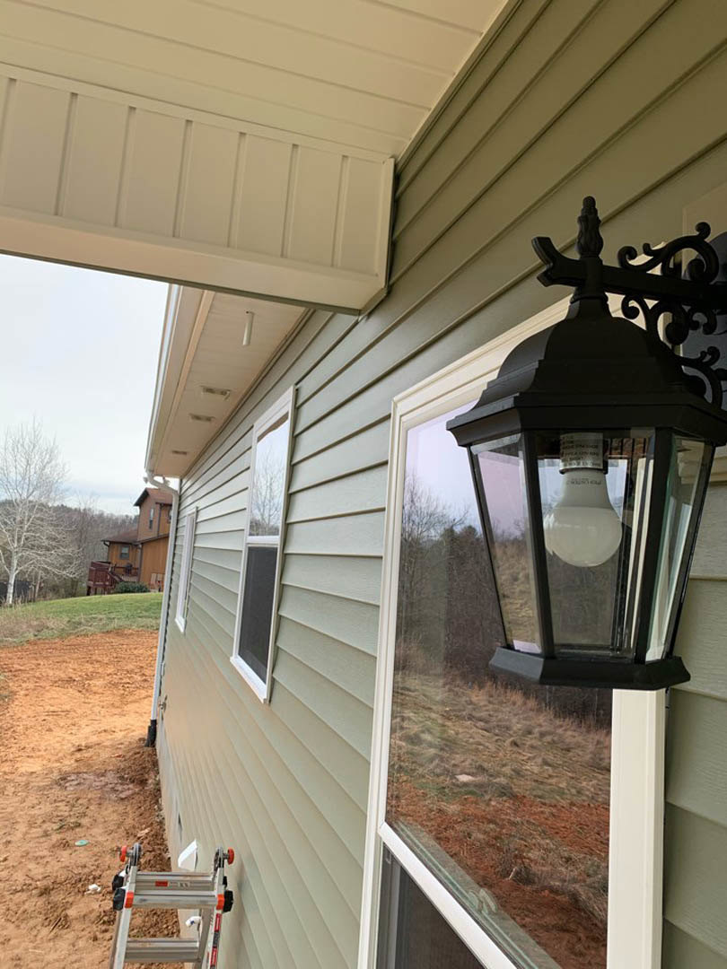 Lantern-style porch light mounted beside window on house exterior, ladder leaning against wall, bare tree and dirt field with excavation hole in foreground, white bottle with black