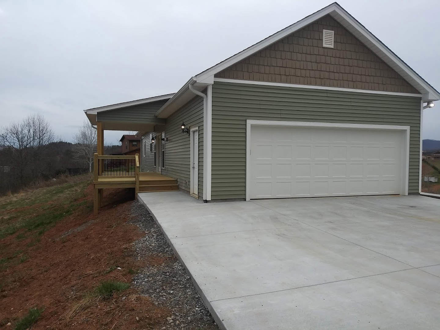 Two-story home with gray siding, attached garage featuring white paneled door, wooden deck with railing on sloped yard, bare tree in front, concrete driveway leading to garage.