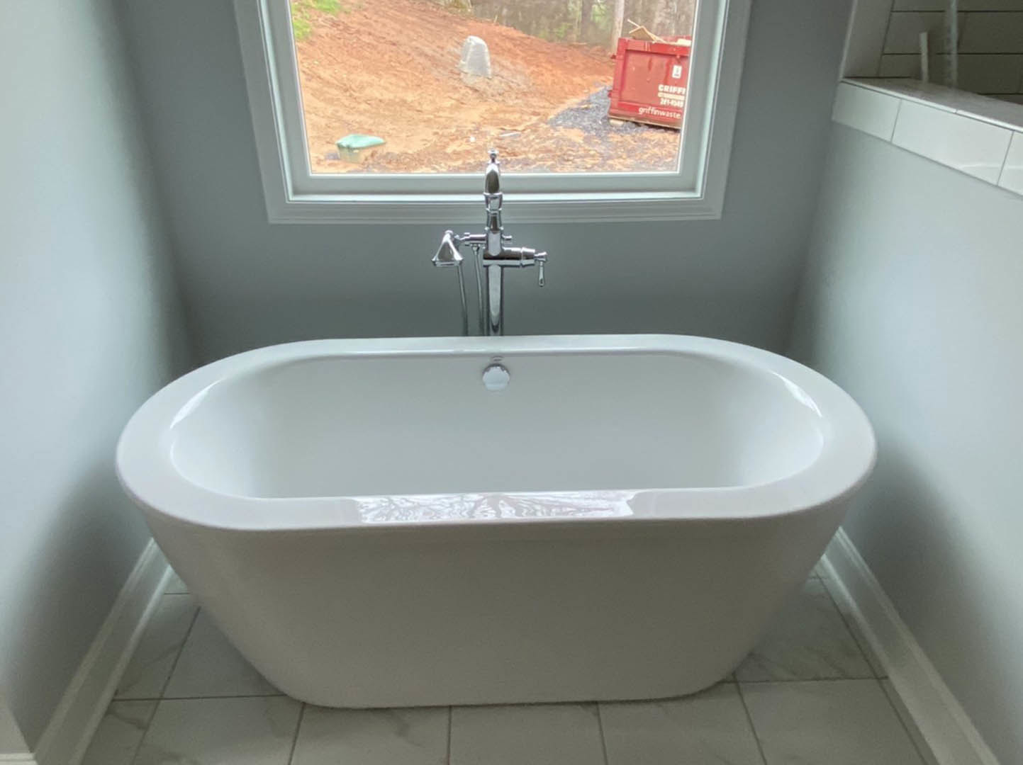 White freestanding bathtub with silver faucet on tiled floor, large window overlooking dirt hill, red container and white object on ground nearby