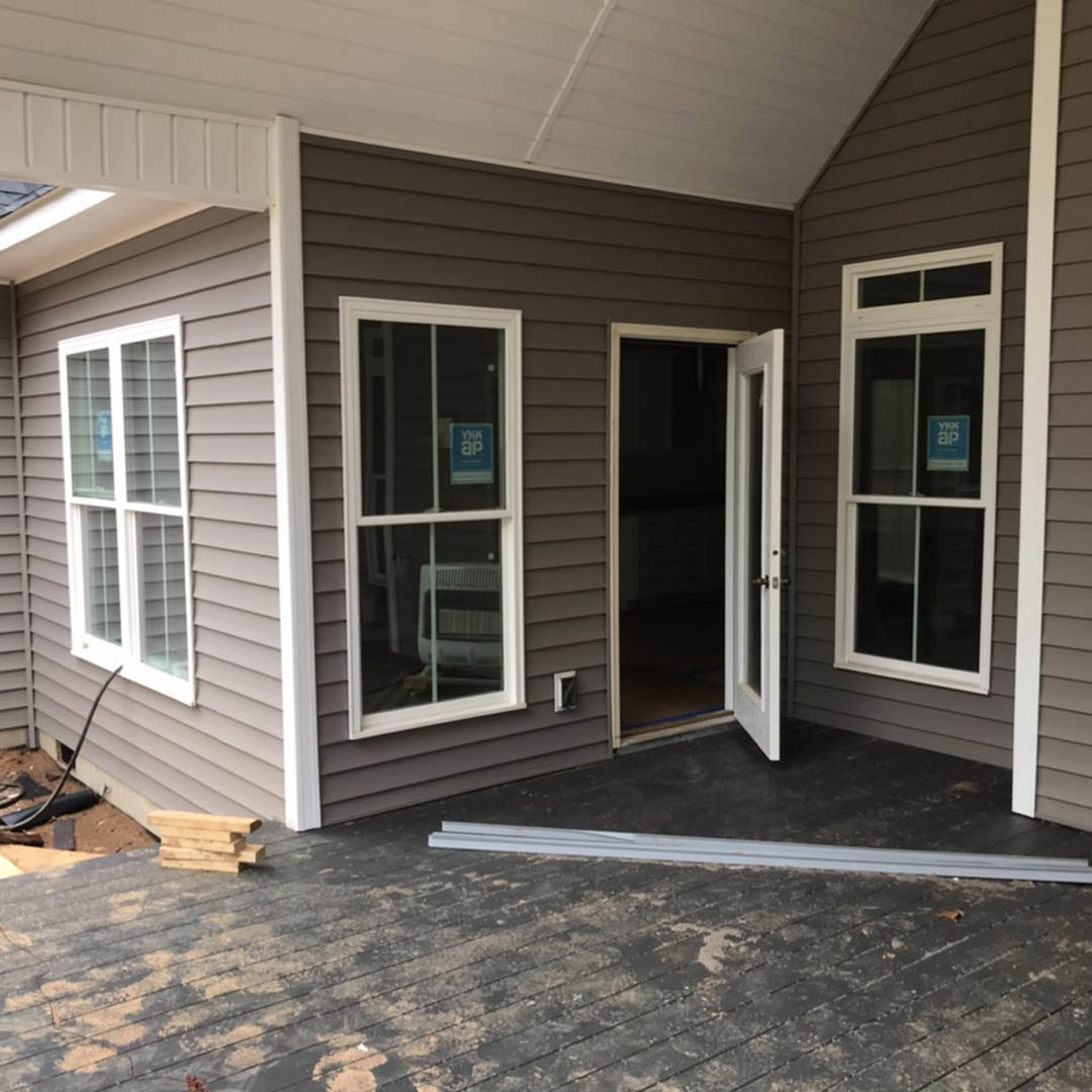 Front porch with white columns, open door, light-colored siding, large window displaying a blue and white sign, outdoor ceiling fixture