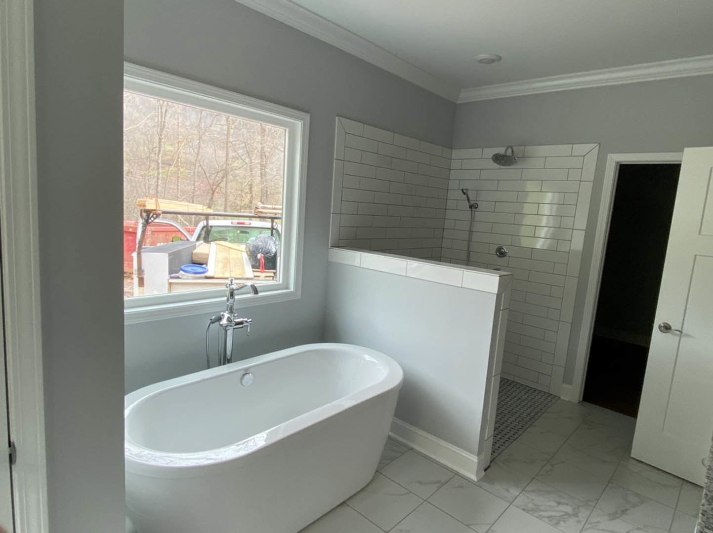 White freestanding bathtub beside glass-enclosed shower with chrome fixtures, gray tile walls, and window overlooking driveway with parked truck and stacked wood