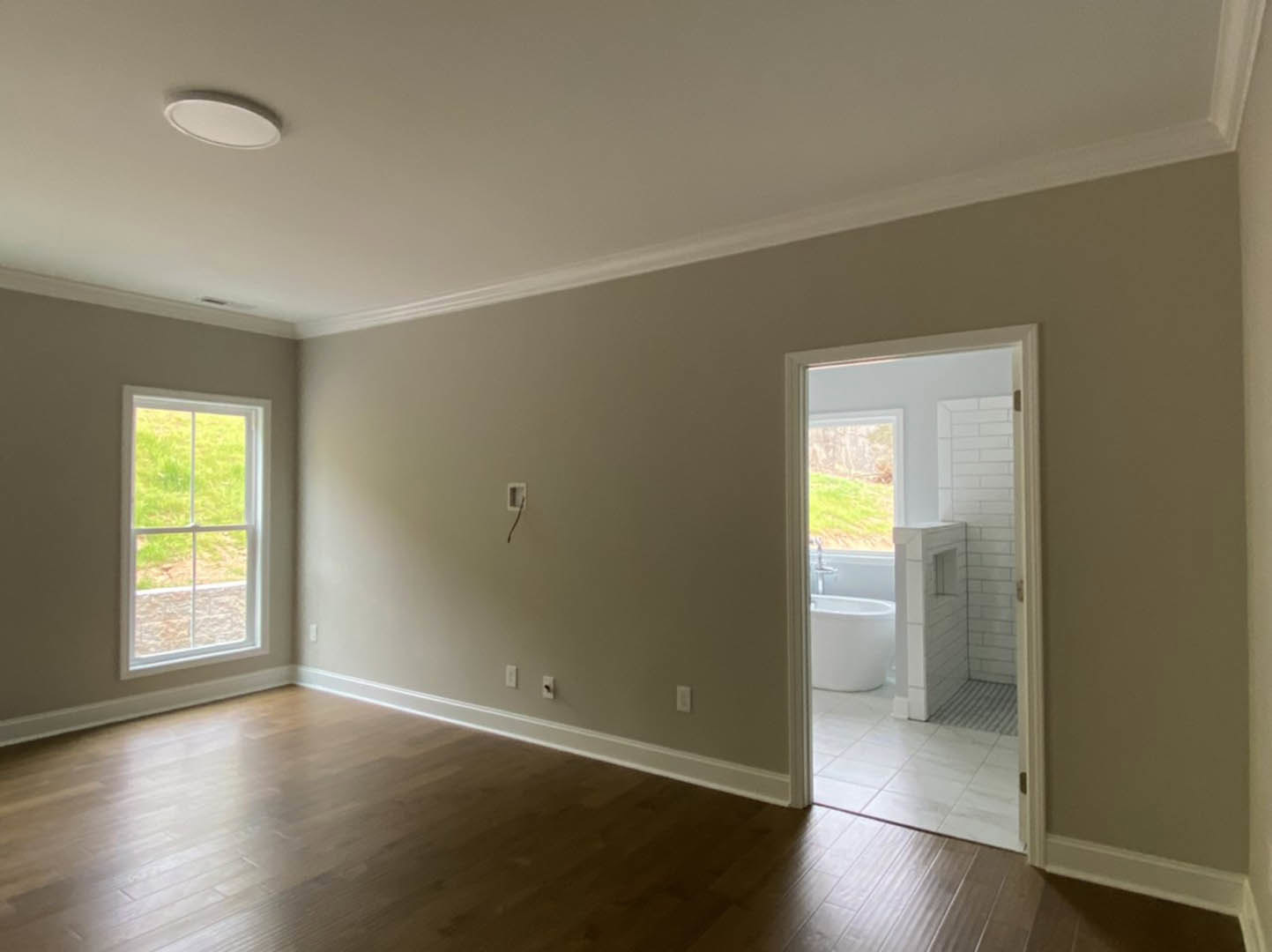 Wood floor and white walls in a room, open door leading to bathroom with white tub and shower, window with white frame, white toilet and bowl visible.