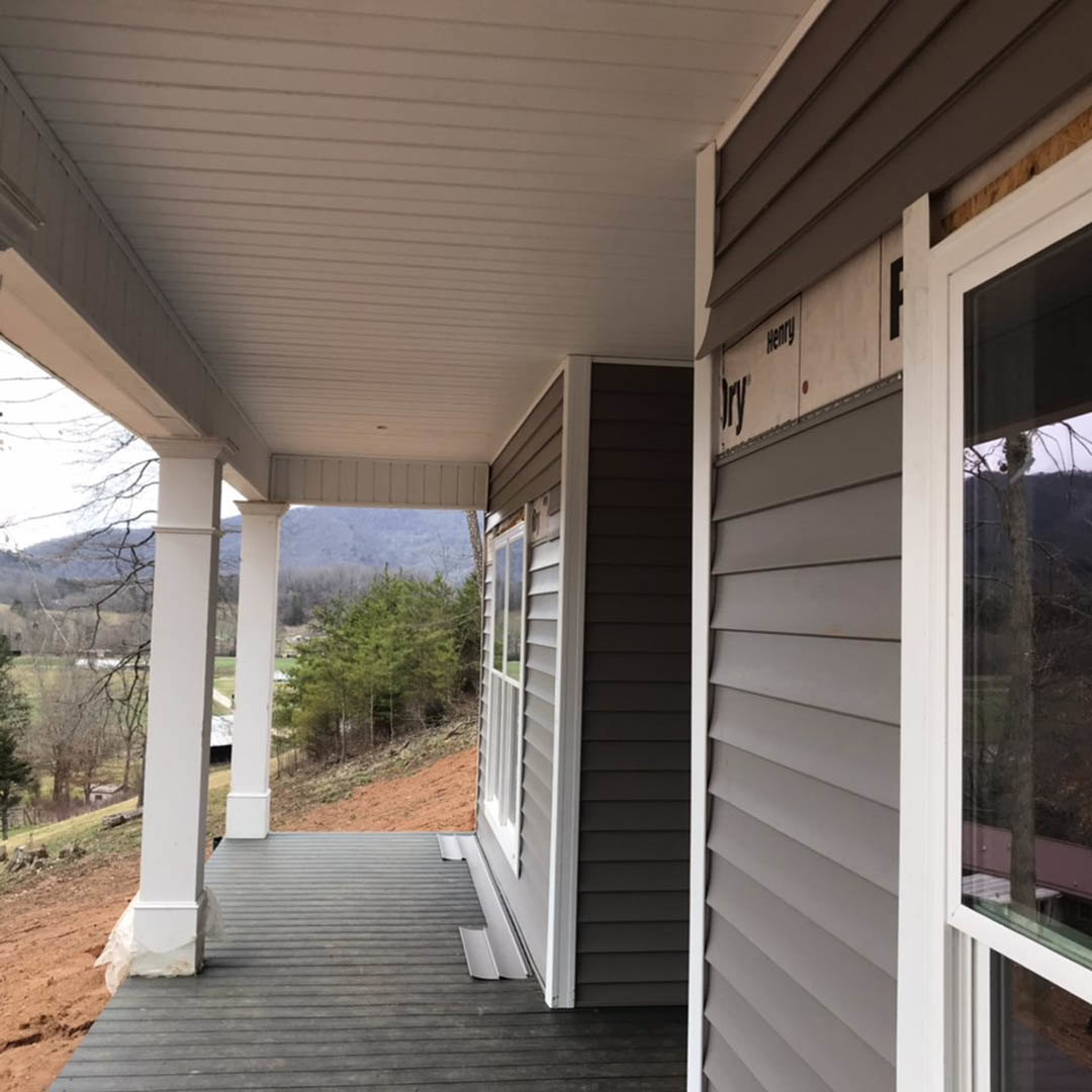 Grey wooden deck with metal railings overlooking a forested hillside, white framed windows and pillars, house exterior facing a valley and dense trees