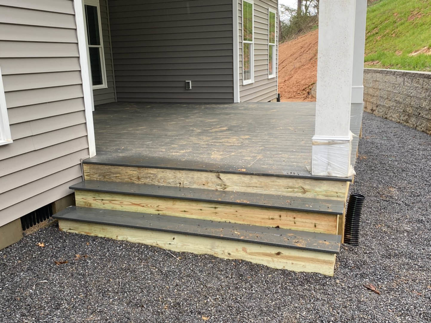 Wooden deck with vertical pillar, stone wall, green grass beyond, and porch area featuring wood plank stairs and dark accent object.
