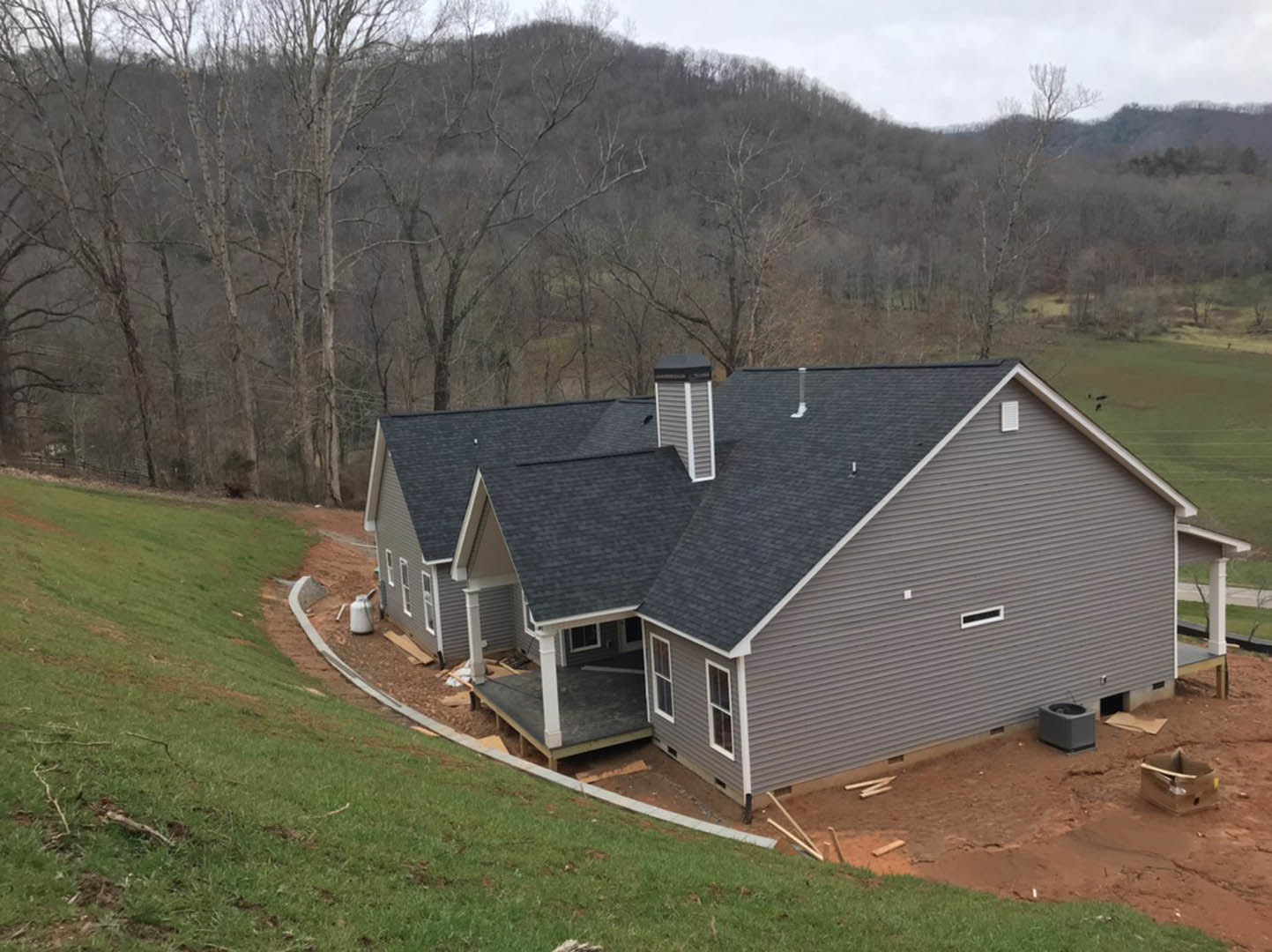 Wood-framed house under construction on a grassy hill, exposed roof trusses, chimney, large windows, surrounded by trees and open sky