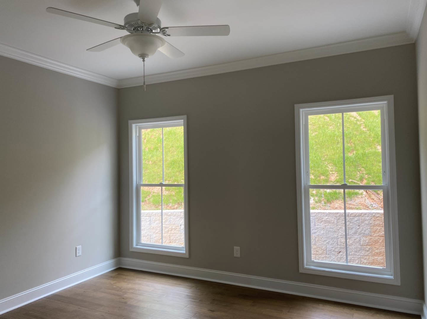 Bedroom with two large windows overlooking a grassy yard, ceiling fan with light fixture, wooden floor, white baseboards, and partial brick accent wall