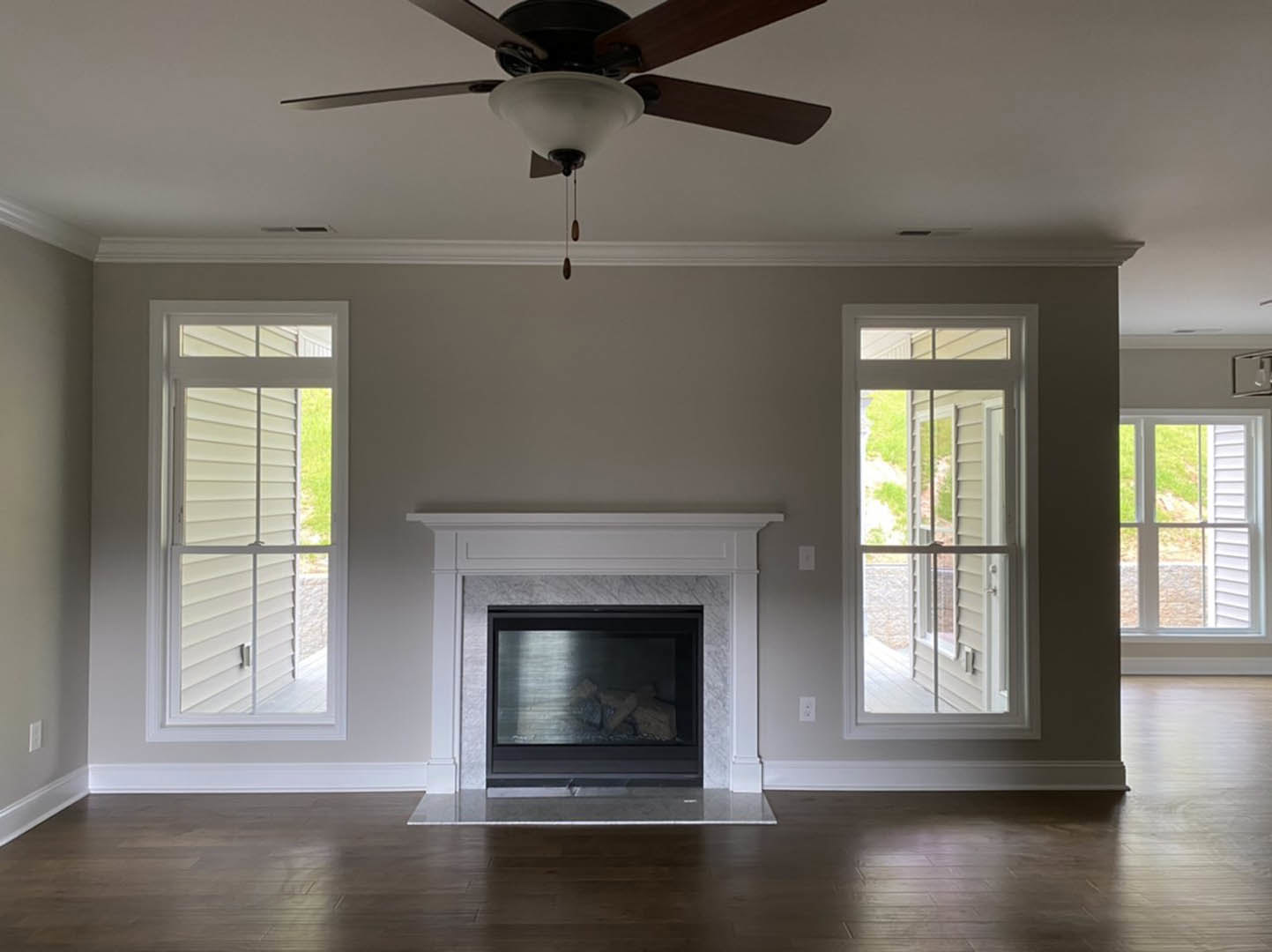 Living room with white-framed fireplace, ceiling fan with light fixture, hardwood floors, and windows with blinds and outdoor view