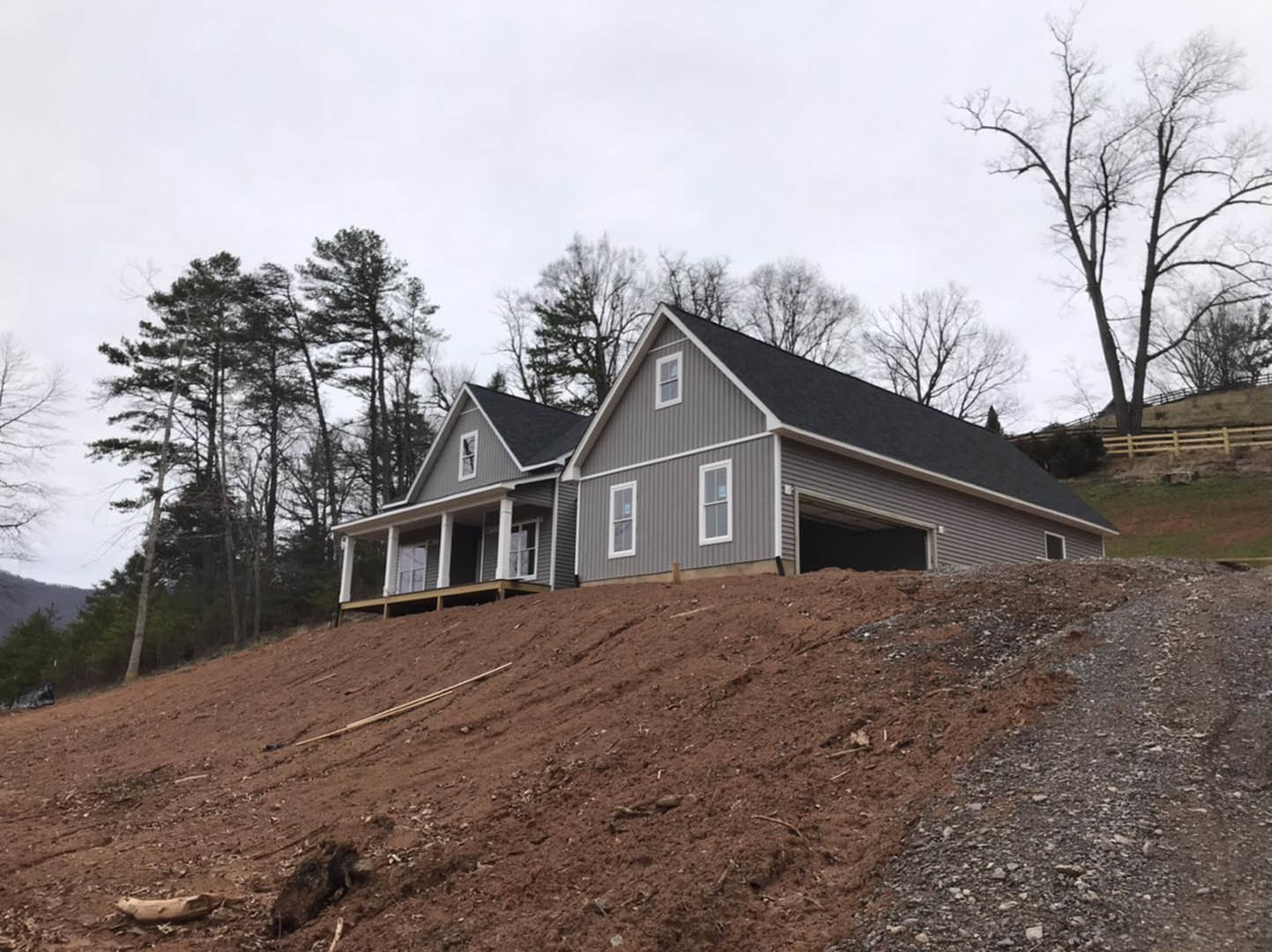 Two-story house with attached garage on grassy hill, leafless tree in foreground, unfinished exterior siding, scattered construction materials, wooded area in background, cloudy