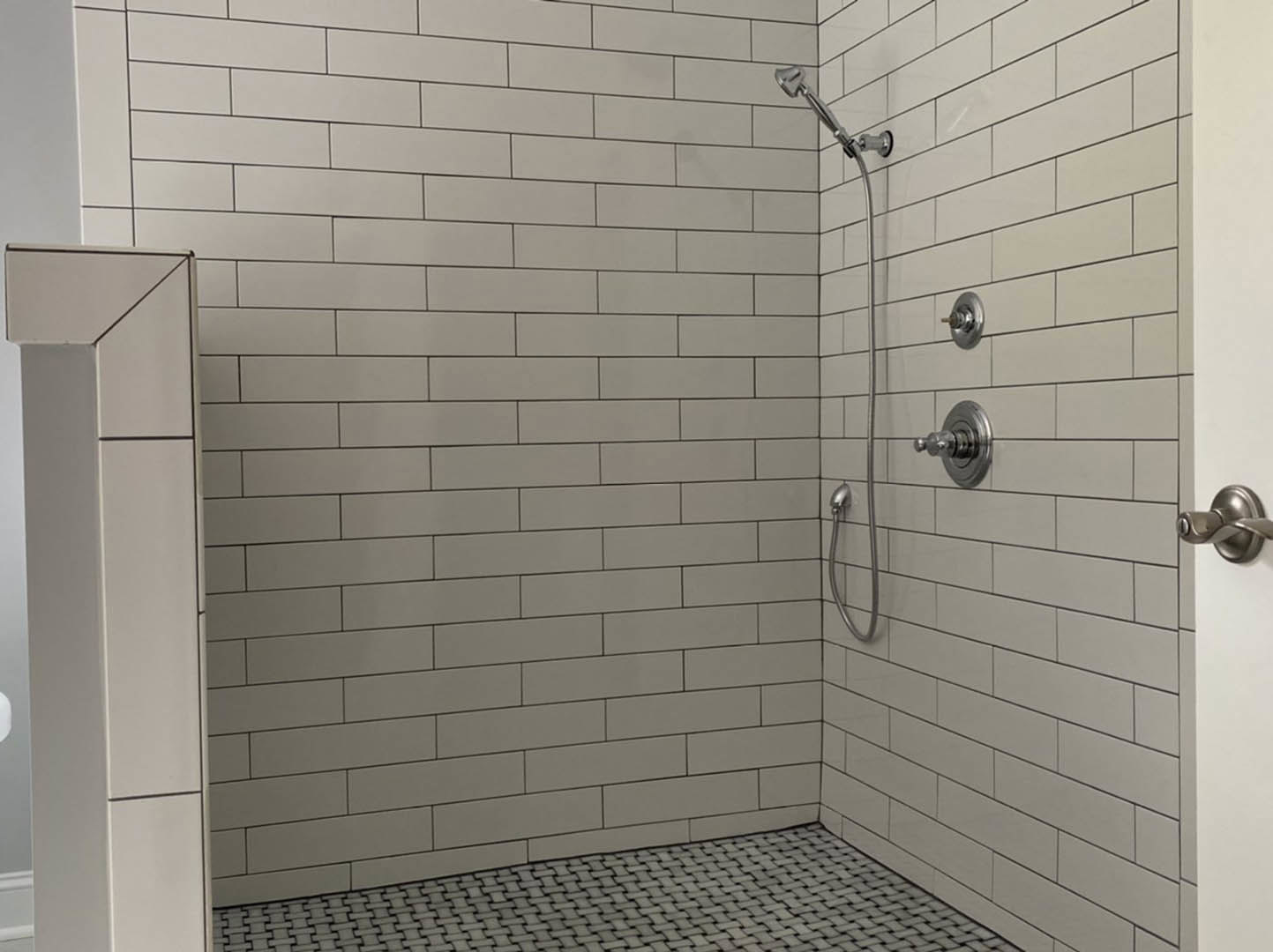 White tiled shower wall with black grout lines, chrome shower head, and glass door with black metal frame