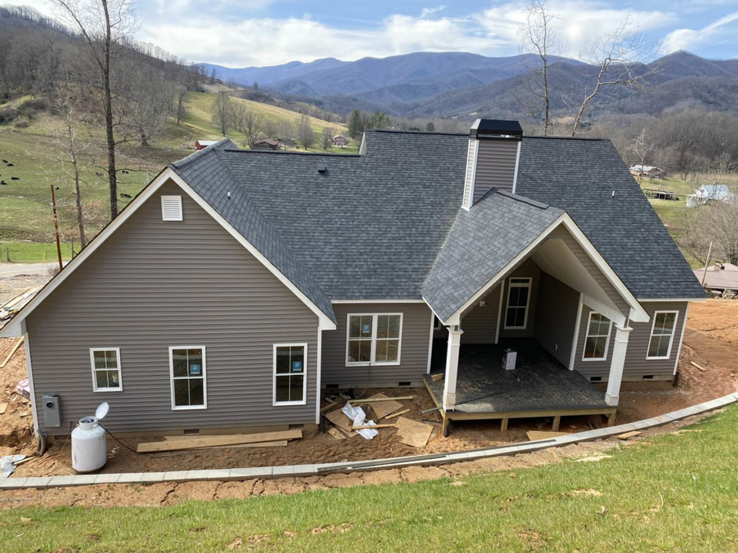 Framed house under construction with exposed plywood walls, large windows, and green lawn, set against a hill and cloudy sky