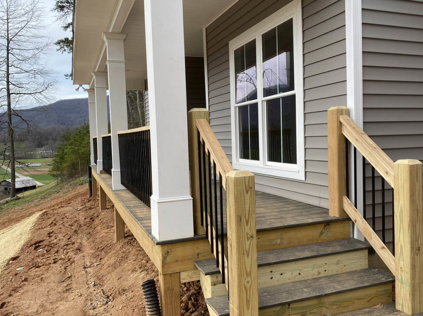 Two-story home with gray siding, wooden porch and stairs, bare tree in front yard, white-trimmed windows, wooden railing and posts on porch