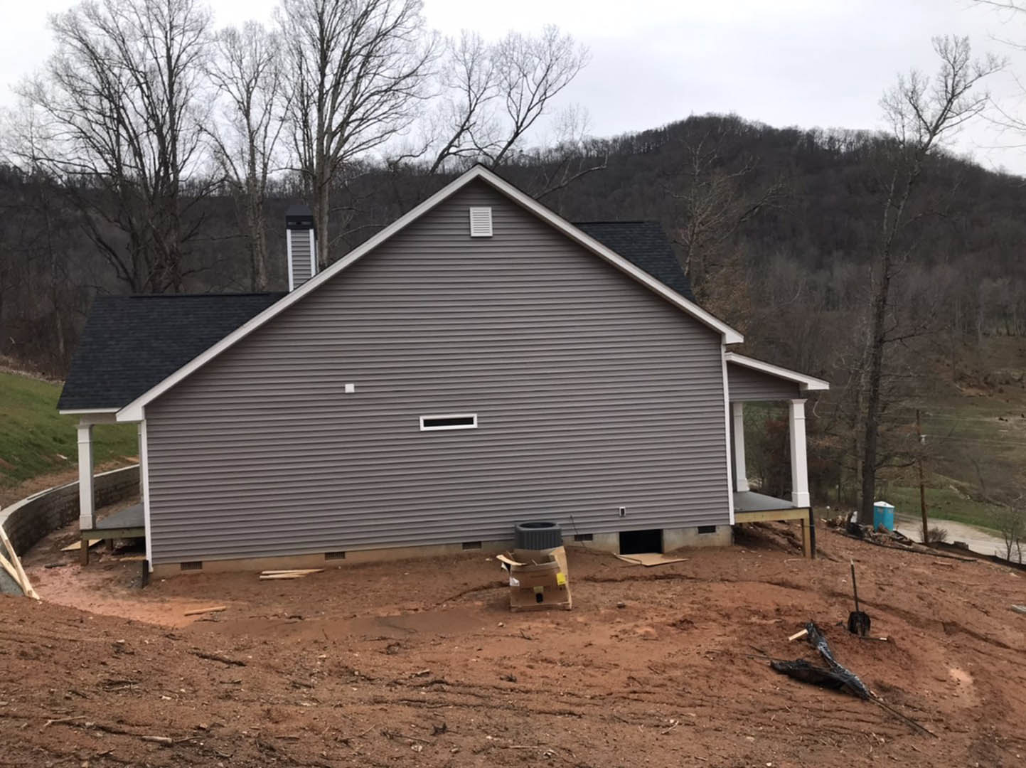 Partially built house with white-framed windows, brick chimney, and unfinished roof surrounded by tall trees; cardboard box and construction materials scattered on bare ground