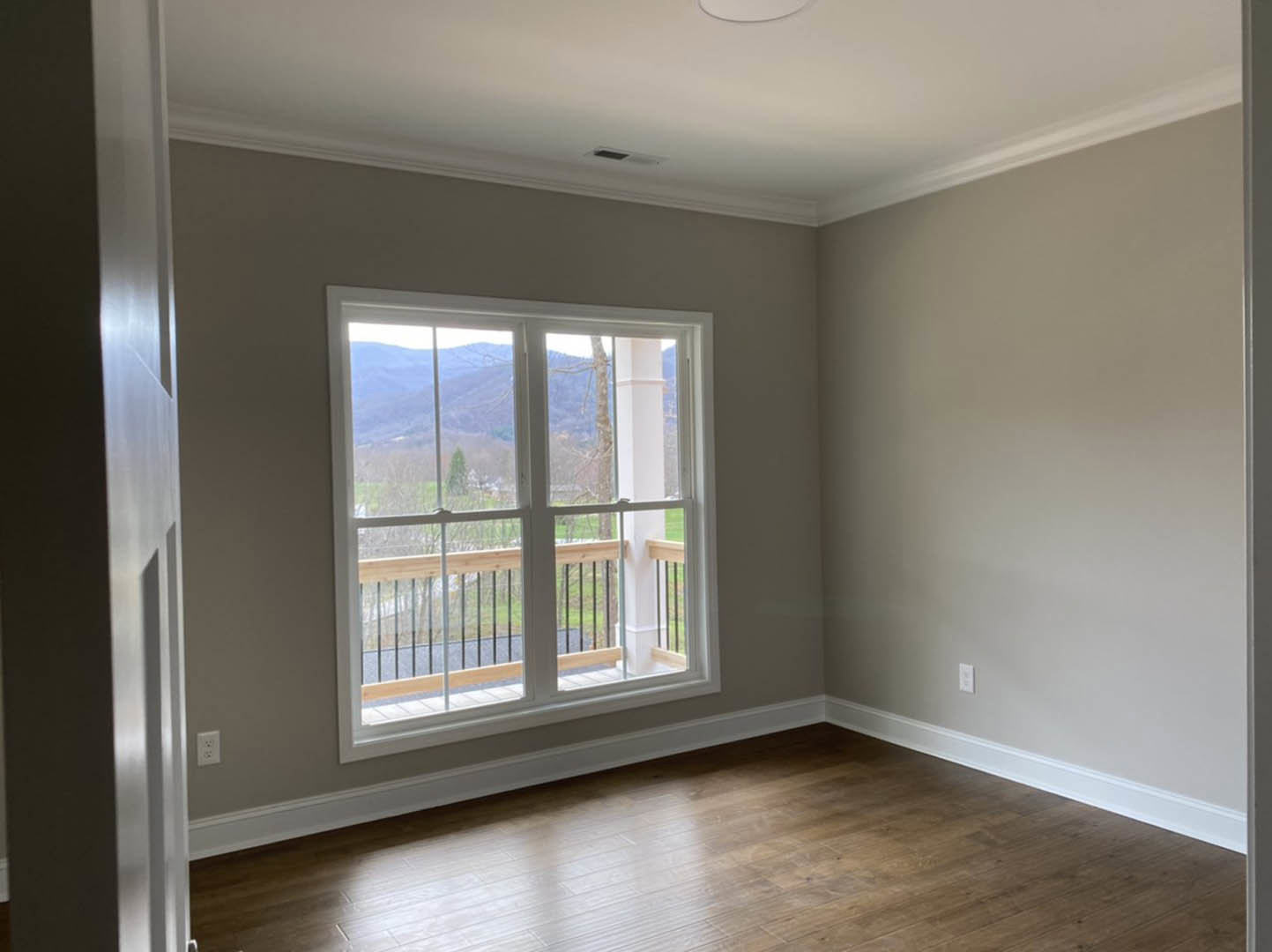 Sunlit room with wood flooring, plaster walls, large window opening to a balcony with wooden railing, mountain and tree views visible outside