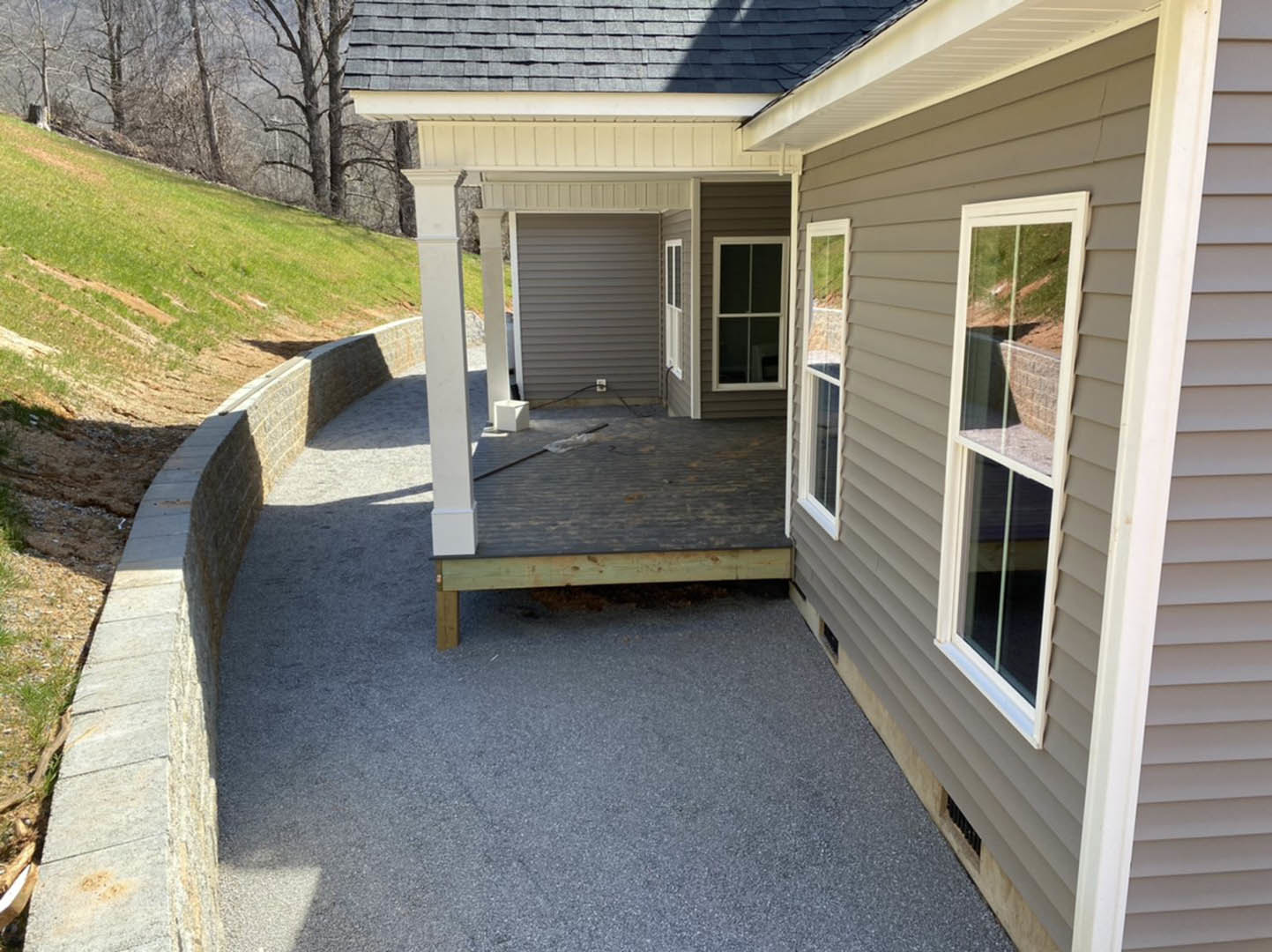 Front porch with white-framed window, stone accent wall, grey siding, shingle roof, and grassy yard with leafless trees
