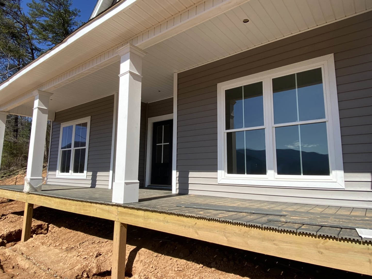 Partially built home with white siding, covered porch, exposed wooden deck, white-framed windows, and sunlight casting shadows on exterior walls.