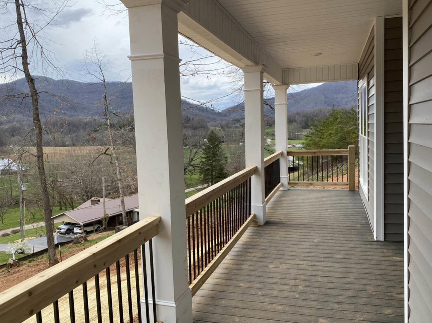 Wooden porch with black metal railings, mountain landscape in background, tree nearby, garage with parked cars visible
