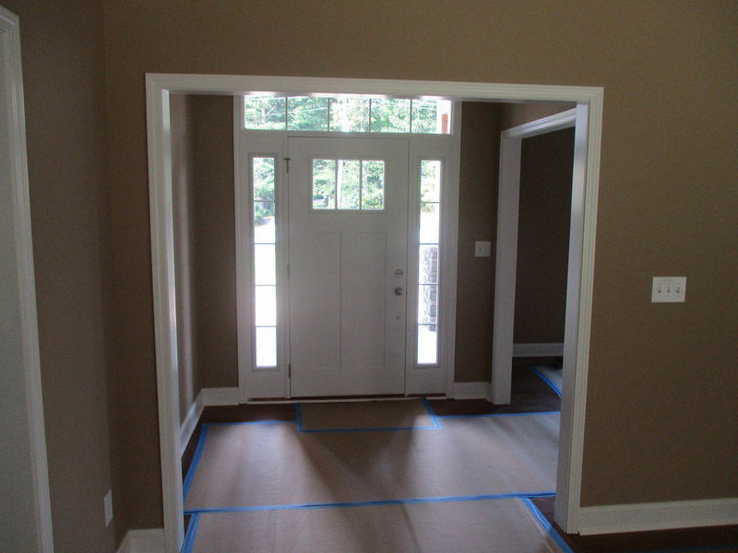 White paneled door with glass windows set in a hallway, blue painter’s tape on trim, white outlet with three switches, dark room beyond doorway, light-colored walls and flooring