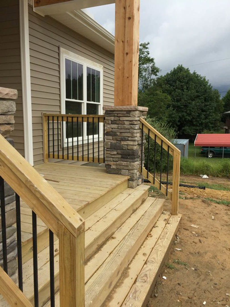 Wooden porch with stone pillars and railing, front stairs leading to entry, close-up window and handrail details, red car roof visible in driveway, surrounded by trees and sky